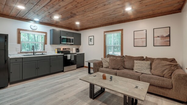 A modern kitchen with grey cabinets, a wooden ceiling, and a dining area with a brown sofa and a coffee table. - Woodsy Cabot Escape in Arkansas Countryside! (Cabot)