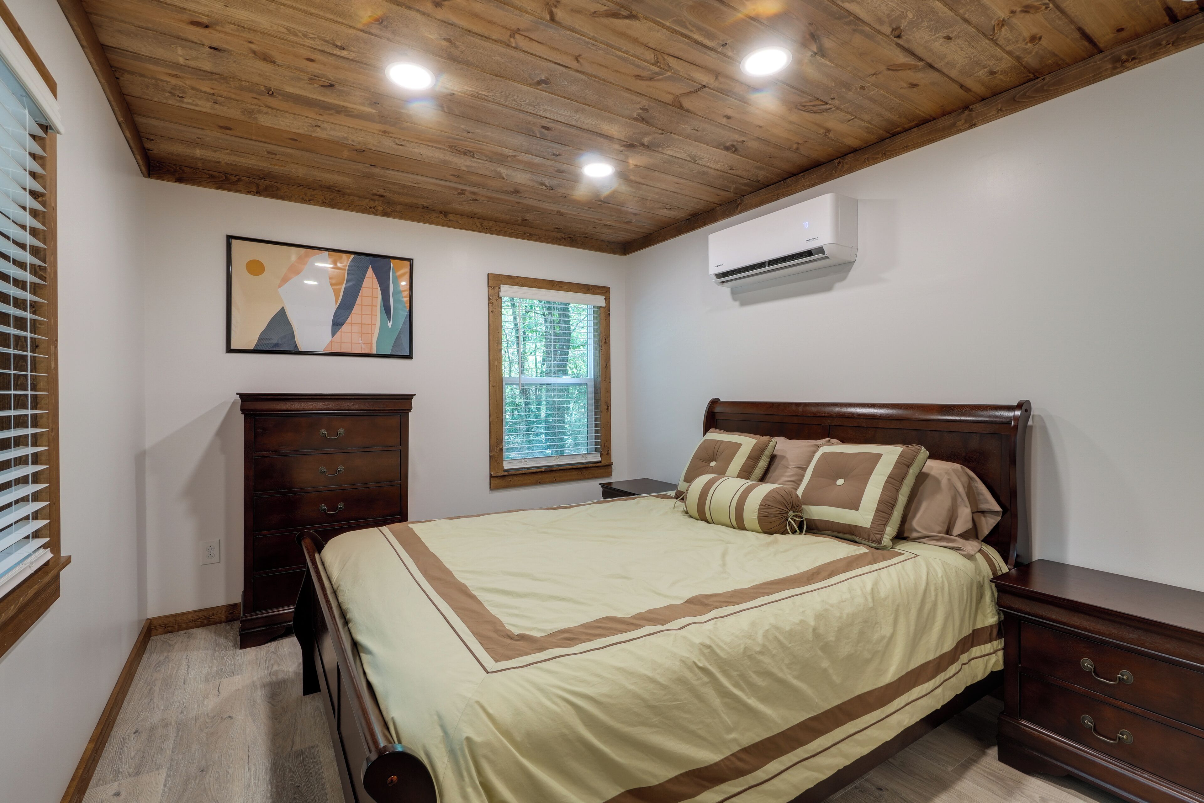A bedroom with a wooden ceiling, a bed with a beige and brown bedspread, a dresser, a nightstand, a window with blinds, and an air conditioning unit.