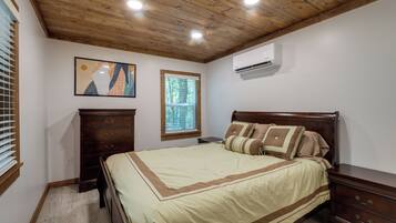 A bedroom with a wooden ceiling, a bed with a beige and brown bedspread, a dresser, a nightstand, a window with blinds, and an air conditioning unit.