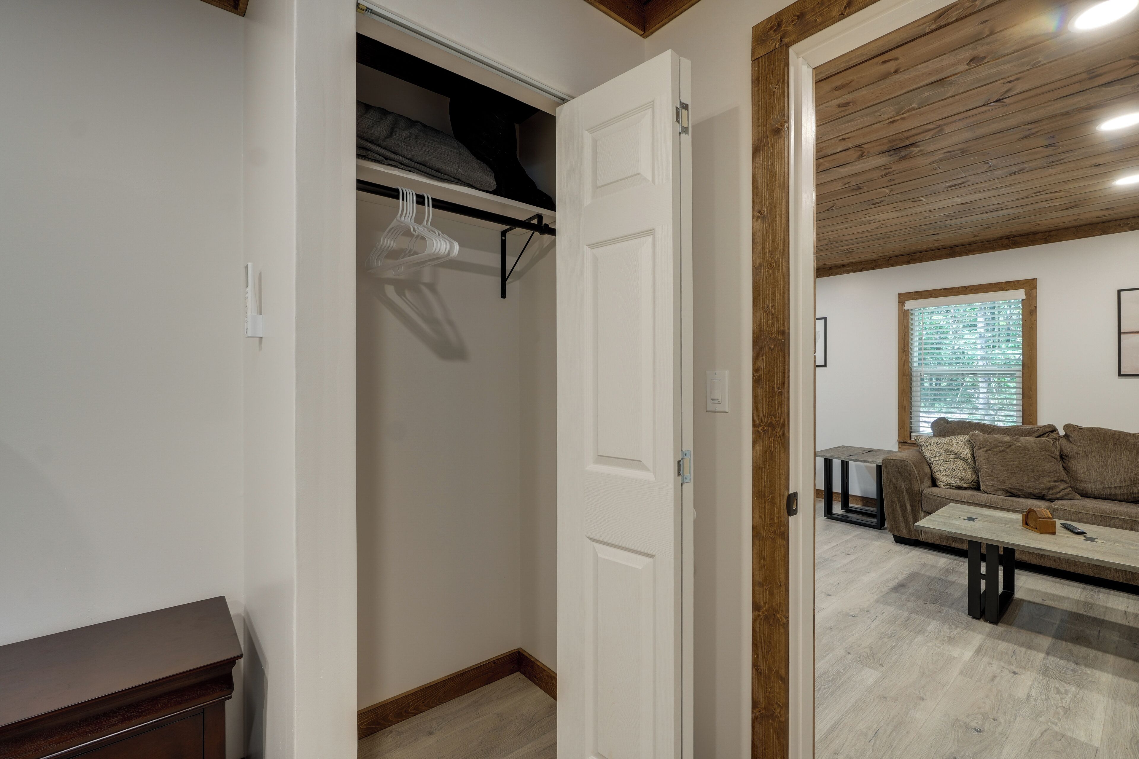 Ein offener Kleiderschrank mit hängender Kleidung, eine Holzbalkendecke, eine Couch und ein Tisch in einem Zimmer mit einem Fenster.