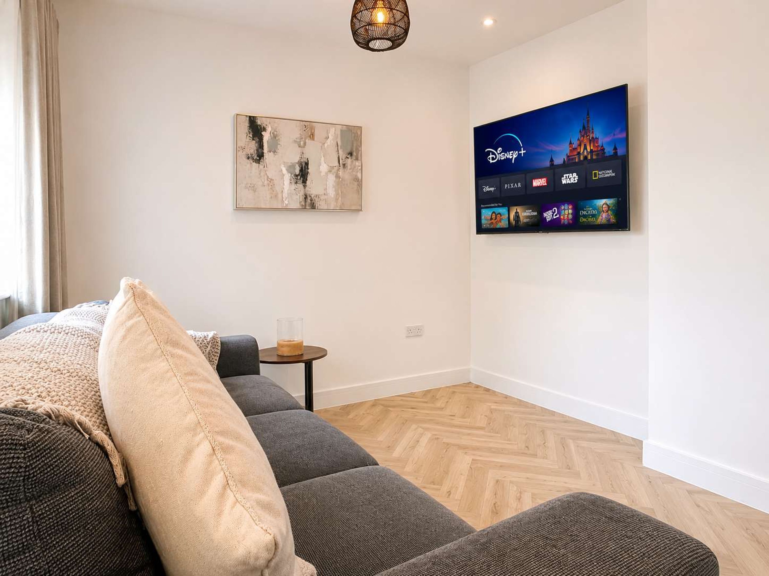 A modern living room with a grey sofa, a coffee table, and a wall-mounted TV displaying Disney+ content.
