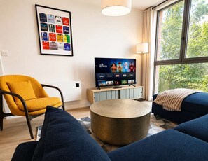 A modern living room with a television displaying Disney and Marvel movies, a framed sports jersey wall art, and a large window with a view of trees. - Luxury 1-apt Old Trafford King Bed (Manchester)