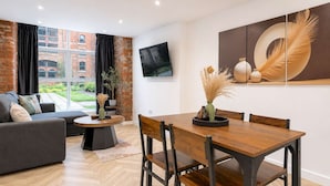 A modern living room with a sofa, a dining table with chairs, a TV mounted on the wall, and a large window with a view of a brick building. - Stylish 1BR - Sleeps 4 - Free Parking - Manchester (Manchester)