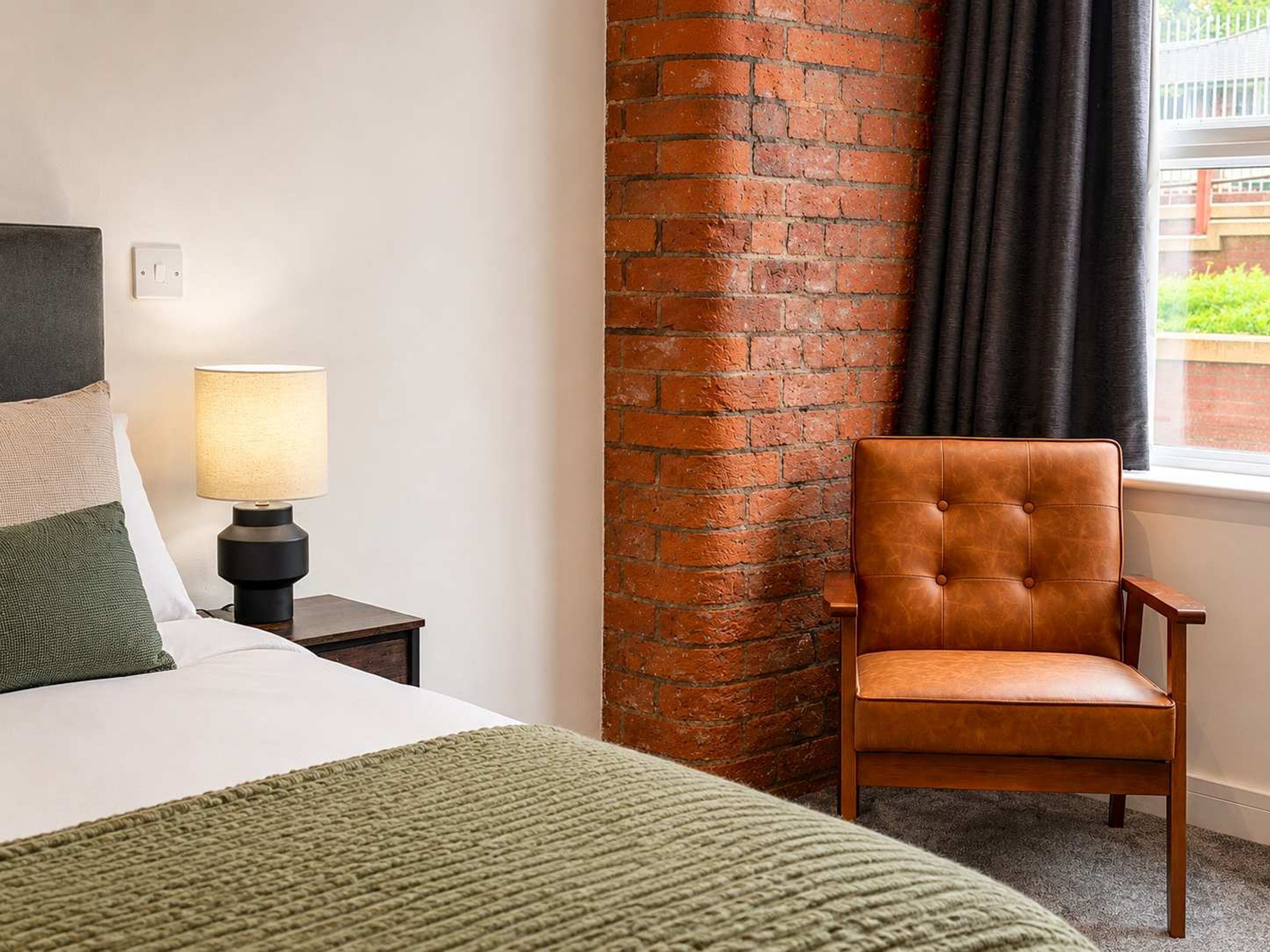 Una camera d'albergo con un letto, una lampada, una poltrona in pelle, un muro in mattoni e una finestra con tende.