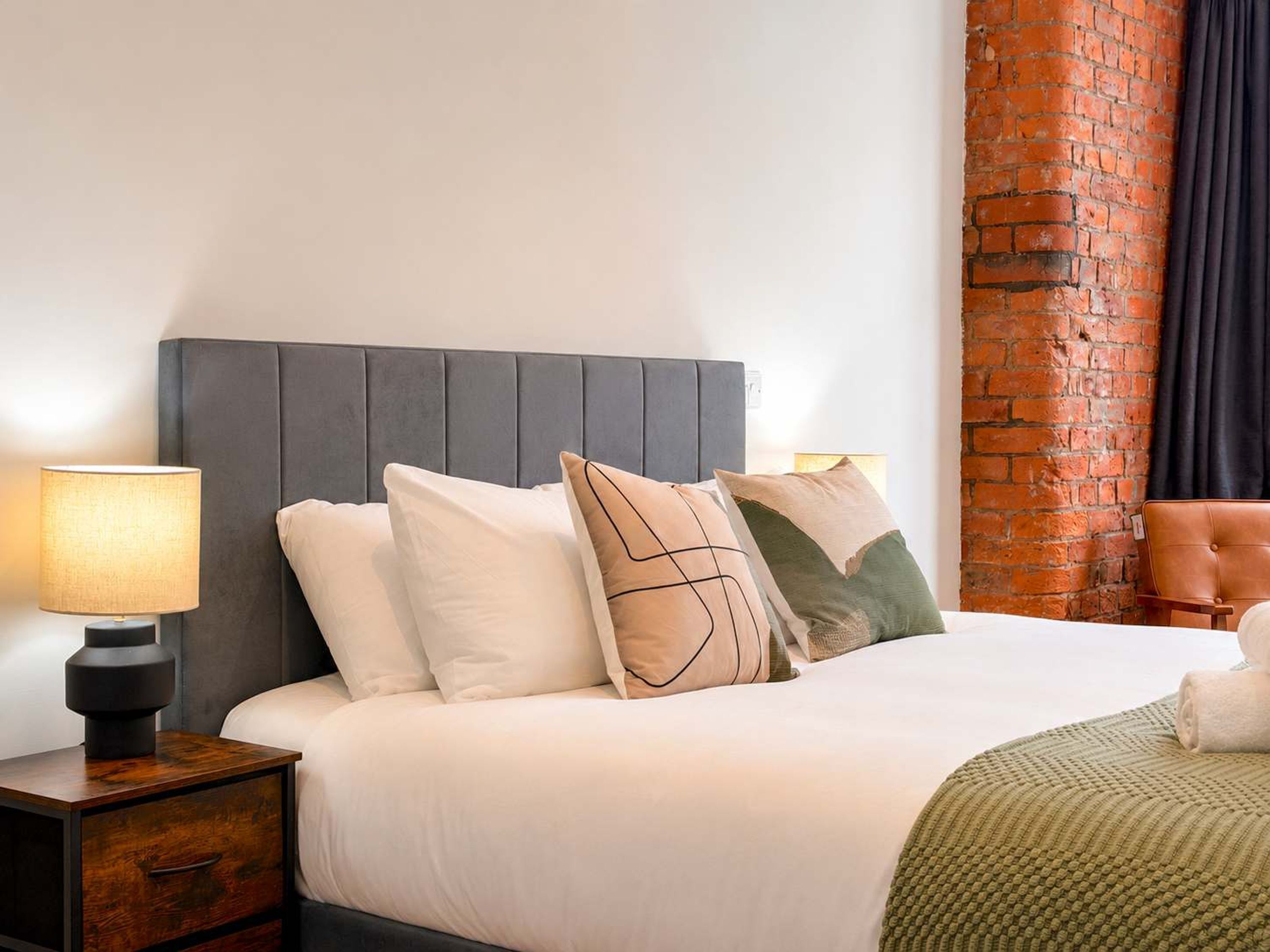 Una cama bien tendida con un cabecero gris, una mesita de noche de madera con cajón y una lámpara.