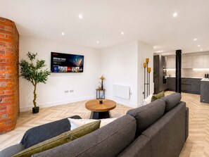 A modern living room with a large flat-screen TV, a brick accent wall, a dining area, and a kitchen in the background. - Sleeps 6 - 2BR Stay - Free Parking - Manchester (Manchester)