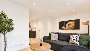 En moderne stue med et rundt træbord, en grå sofa med mønstrede puder, et stort abstrakt maleri og en potteplante.