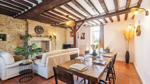 Un intérieur rustique avec des poutres apparentes, une cheminée en pierre, une table à manger dressée pour quatre personnes et un canapé blanc.
