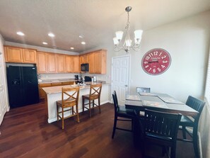 Una cocina con armarios de madera, un refrigerador negro, un microondas, una araña y un comedor con una mesa y sillas.