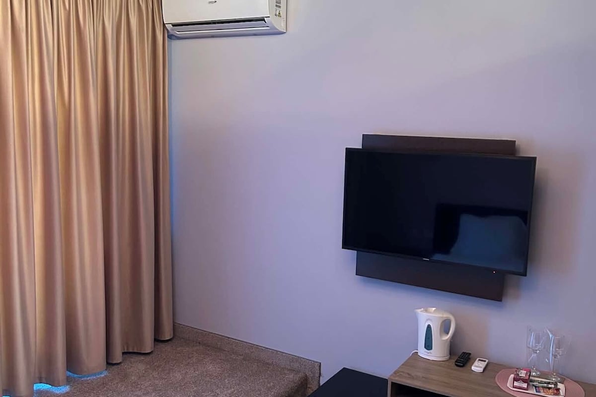 Hotelkamer met een flatscreen-tv aan de muur, een klein bureau met waterkoker, afstandsbediening en een glas water. Een bed met een witte handdoek en een vloerkleed.