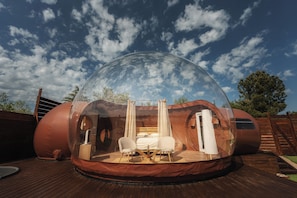 A transparent dome structure with a bed, two chairs, and a small table, set on a wooden deck.