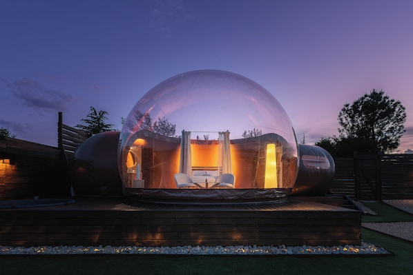 A transparent dome structure with a seating area inside, surrounded by a wooden deck and a fence.
