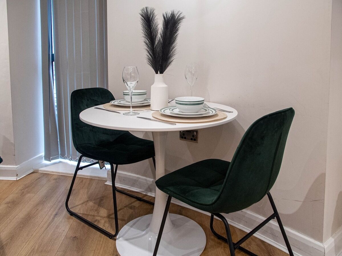 Een eetgedeelte met een ronde tafel, twee donkergroene stoelen, witte borden en een vaas met veertjes.