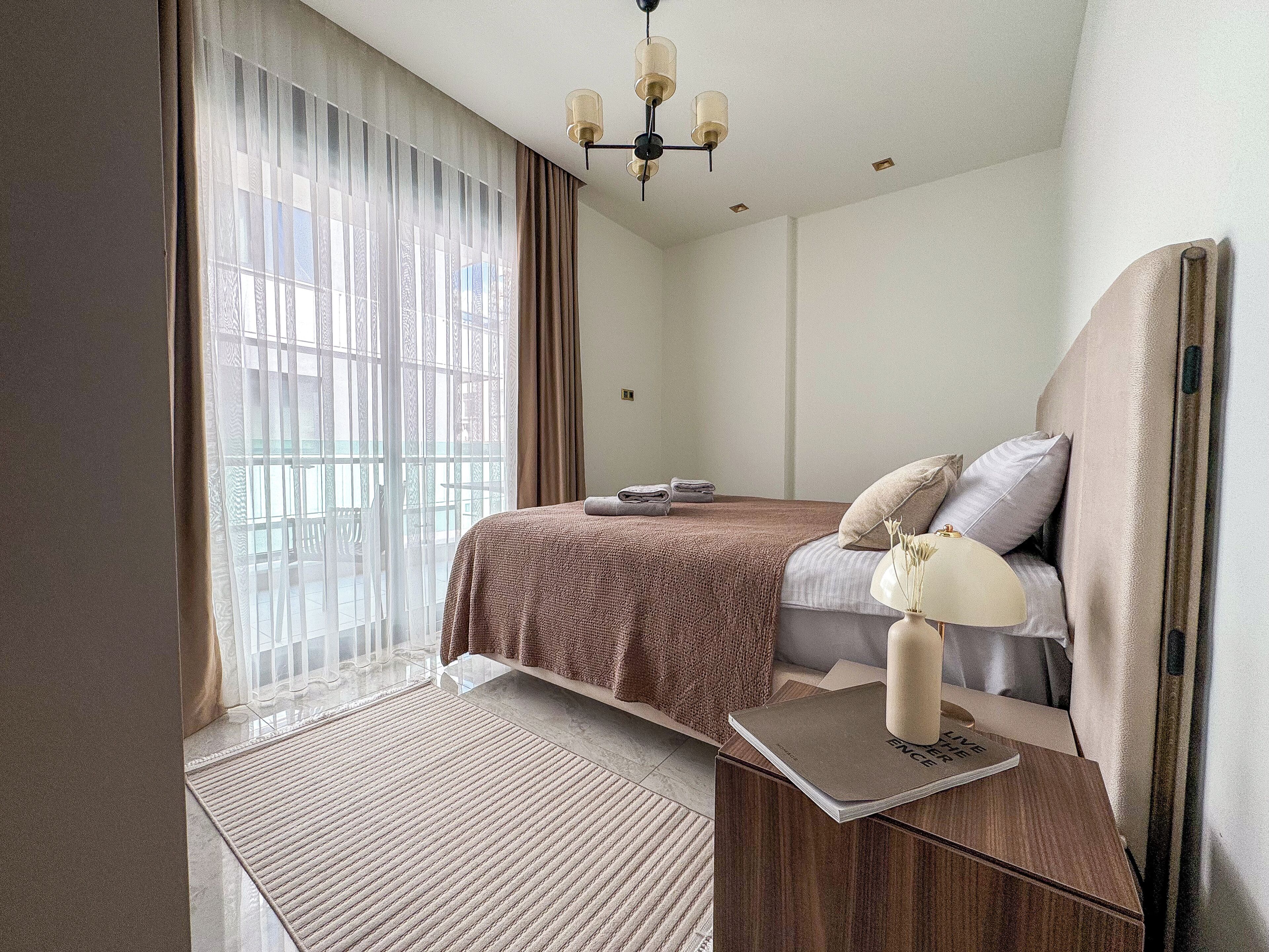Quarto com cama, mesa de cabeceira, abajur e vista para o exterior pela janela.
