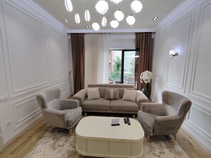 Un salon avec un canapé beige, deux fauteuils, un pouf blanc et une vue sur l’extérieur grâce à une grande fenêtre.