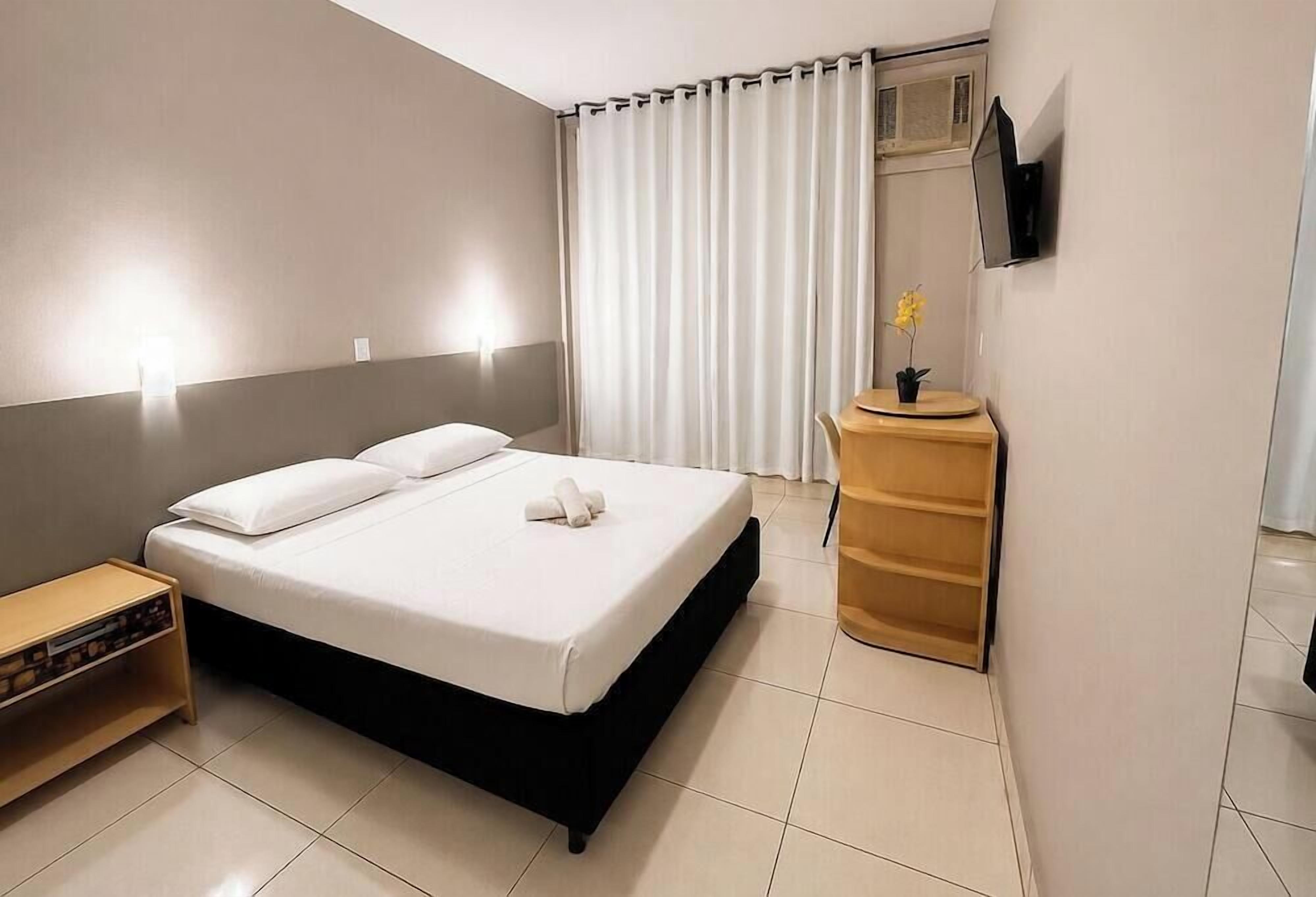 A hotel room with a bed, bedside table, and a small wooden cabinet.