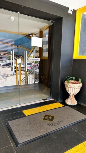 A hotel entrance with a glass door, a yellow tactile paving strip, a doormat with a logo, and a potted plant. - Riviera Hotel Brasilia (Brasília)