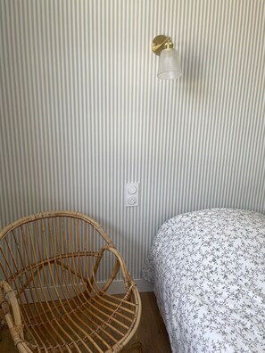 Ein Zimmer mit gestreifter Tapete, einem Korbstuhl, einem Bett mit floralem Überwurf und einer wandmontierten Leuchte.