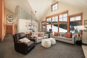 Ein geräumiges Wohnzimmer mit hoher Decke, großen Fenstern mit Blick auf eine verschneite Landschaft, einem Ledersessel, einem grauen Sofa mit vielen Kissen und einem kleinen Tisch mit einer Lampe.