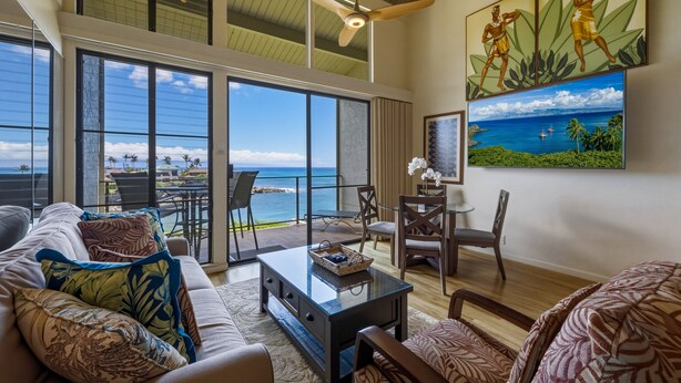 A spacious living area with a large window overlooking the ocean, a dining area with a view, and tropical-themed artwork. - Ocean View 2BR Loft Near Napili Beach - Unit C34 (Lahaina)