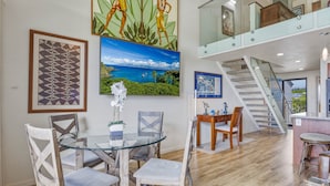 A spacious living area with a glass dining table, wooden chairs, and a staircase leading to an upper level. - Ocean View 2BR Loft Near Napili Beach - Unit C34 (Lahaina)