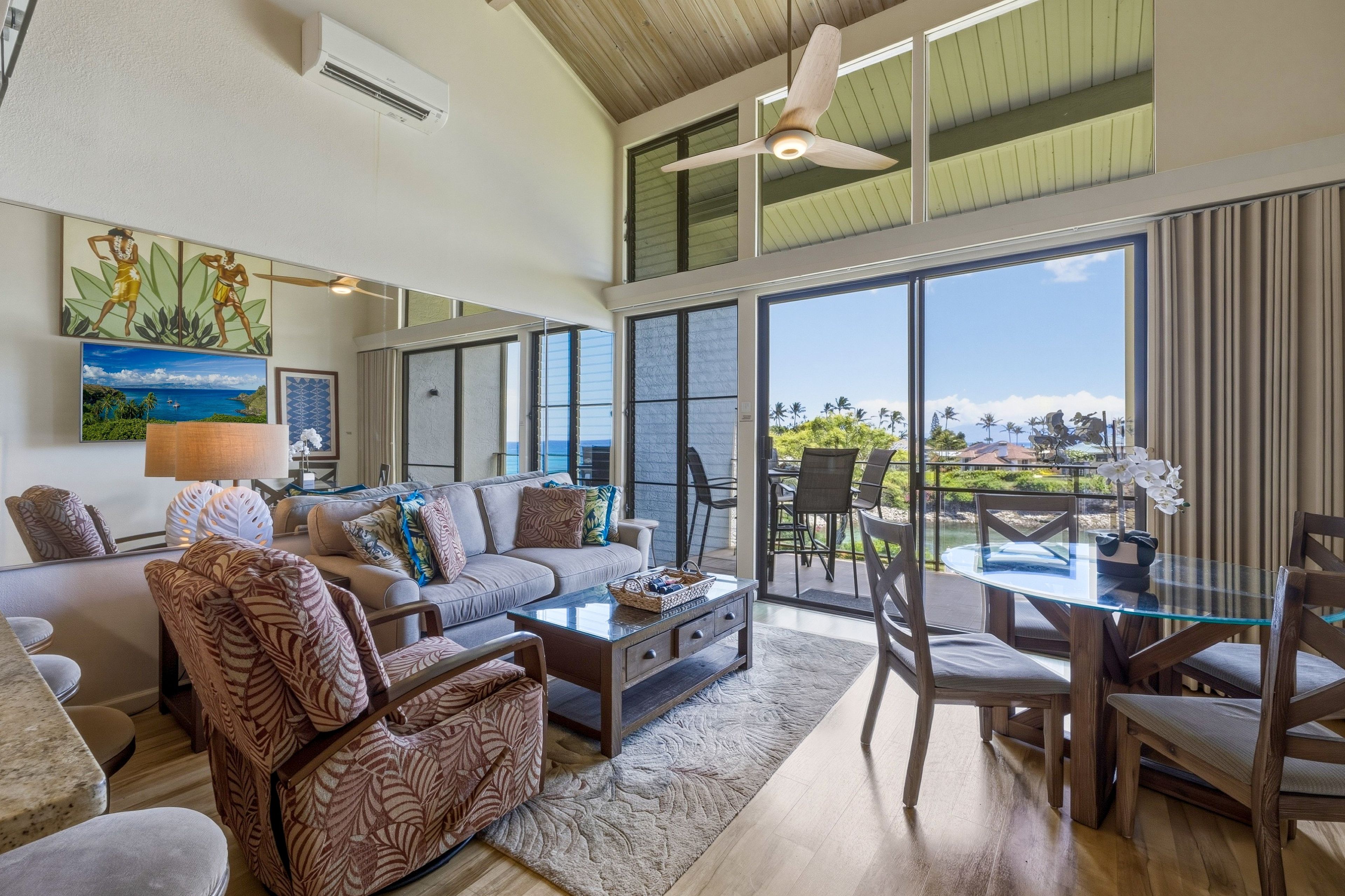 Ein geräumiges Wohnzimmer mit einer großen Couch, einem Couchtisch und einem Essbereich mit Stühlen und einem Glastisch. Ein Deckenventilator, ein großes Fenster mit Meerblick und Kunstwerke an den Wänden sind ebenfalls vorhanden.