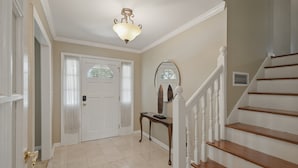Käytävä, jossa on valkoinen ovi, puinen konsolipöytä, peili ja valkoisilla kaiteilla varustetut portaat.
