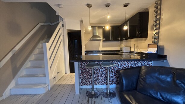A modern kitchen with black cabinets, a central island, and a staircase leading to another level. - The Secluded Corner Wimbledon Chase With Parking (London)