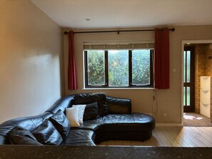 A room with a black leather sofa, a window with red curtains, and a stone wall. - The Secluded Corner Wimbledon Chase With Parking (London)