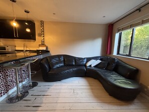 A living room with a black leather sofa, a kitchen area with a microwave, and a window with a view of trees. - The Secluded Corner Wimbledon Chase With Parking (London)