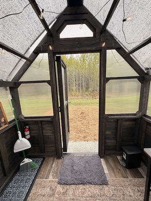A greenhouse with a wooden interior, a door leading outside, a broom, a fire extinguisher, and a light fixture.