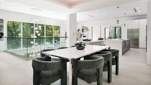 A modern kitchen with a central island, a dining area with a white table and grey chairs, and a glass railing overlooking greenery. - Luxury Brickell Home, Rooftop Pool, BBQ, City Lights (Miami)