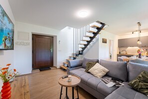 Ein modernes Wohnzimmer mit einem grauen Sofa, einem kleinen runden Holztisch, einer Treppe mit Holzstufen und schwarzen Geländern sowie einer Küchenzeile im Hintergrund.