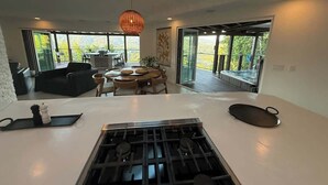 A modern kitchen with a central island, dining area with round table, and large windows offering a view of the outdoors.