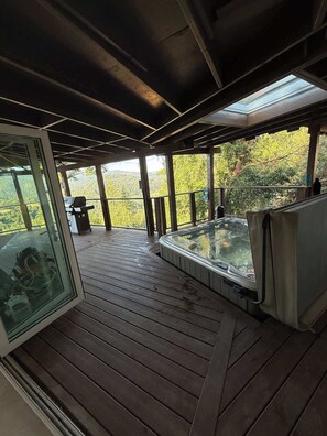 Une terrasse en bois dotée d’un jacuzzi et d’un espace clos par des parois de verre, offrant une vue sur un paysage boisé.