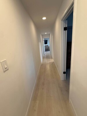 Un couloir étroit, doté d’un plancher en bois et de murs blancs, mène à une pièce où se trouvent un lit et une commode.