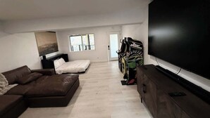 A modern living room with a flat-screen TV, a brown leather sofa, a gaming console, and a window providing natural light.