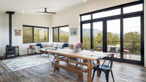 Amplia sala de estar con una mesa de madera, bancos, un sofá y grandes ventanales con vistas al exterior.