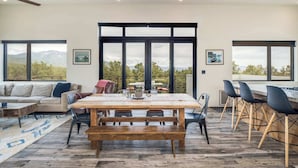 Un salón amplio con una gran mesa de madera, sillas negras, un sofá y vistas a montañas a través de amplios ventanales.