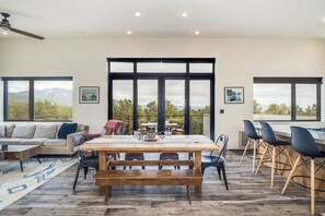 Ein geräumiges Wohnzimmer mit einem großen Holztisch, schwarzen Stühlen, einer Couch und Bergblick durch große Fenster.