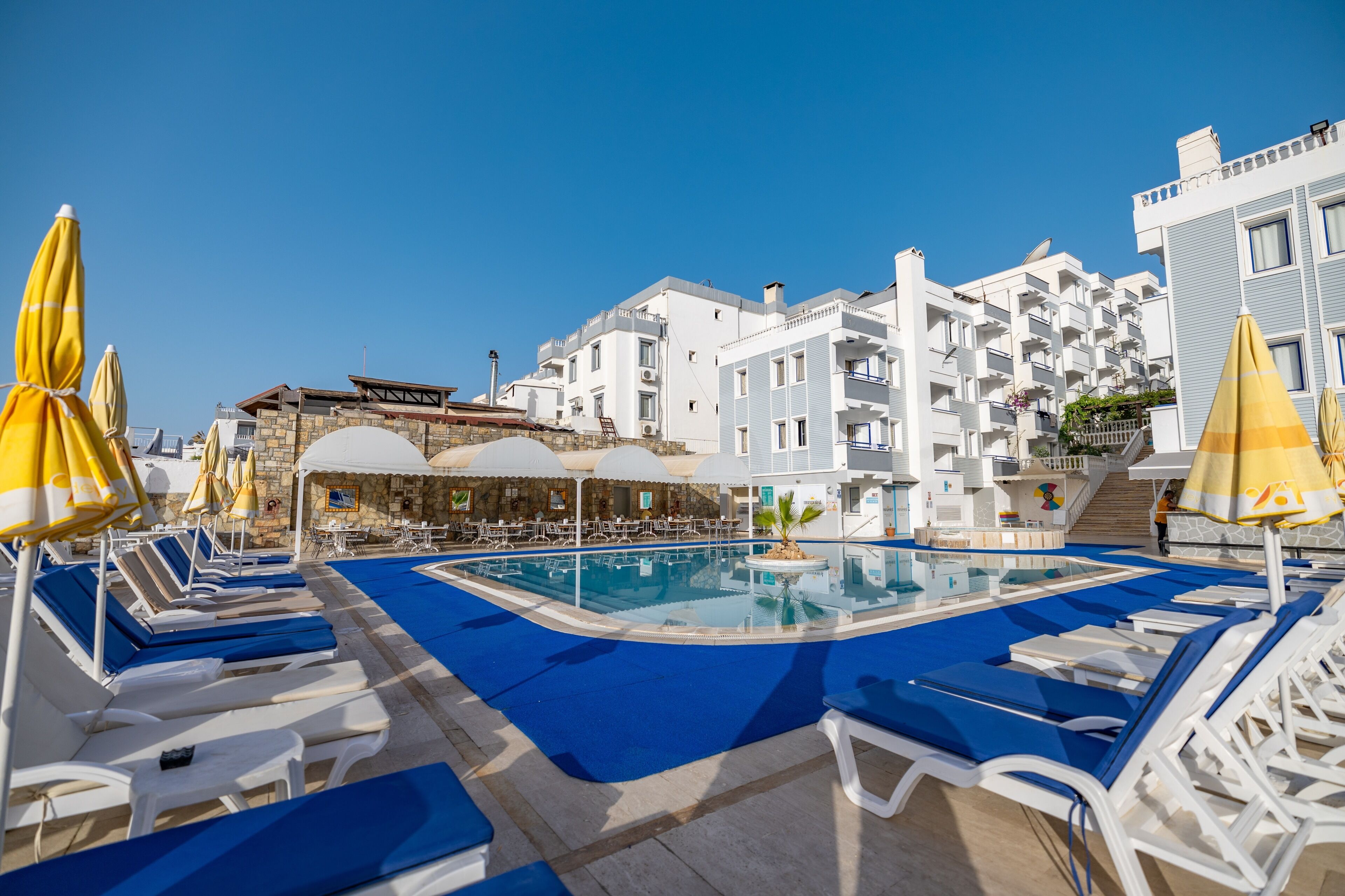 A pool area with lounge chairs and umbrellas, a clear blue sky, and white buildings in the background.