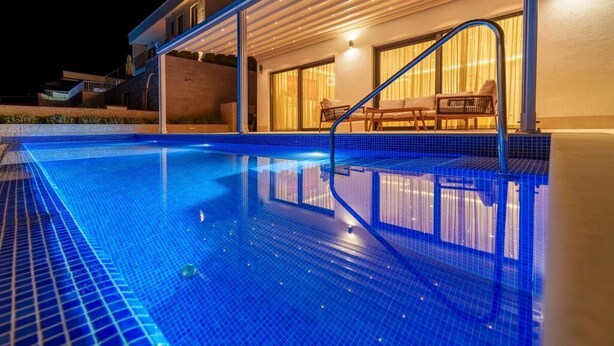 En swimmingpool med blå mosaikgulv, et overdækket siddeområde med stole, og en bygning med store vinduer.