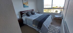 Une chambre à coucher avec un lit, une table de chevet, une chaise, une fenêtre donnant sur un beau paysage et un tableau accroché au mur.