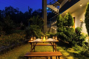 Una mesa de comedor al aire libre con velas, rodeada de follaje y una escalera.