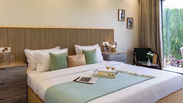 A hotel room with a large bed, bedside tables, a chair, and a view of greenery through the window. - ELIVAAS Aranya Library Villas With Pool (Fatehpur Range)