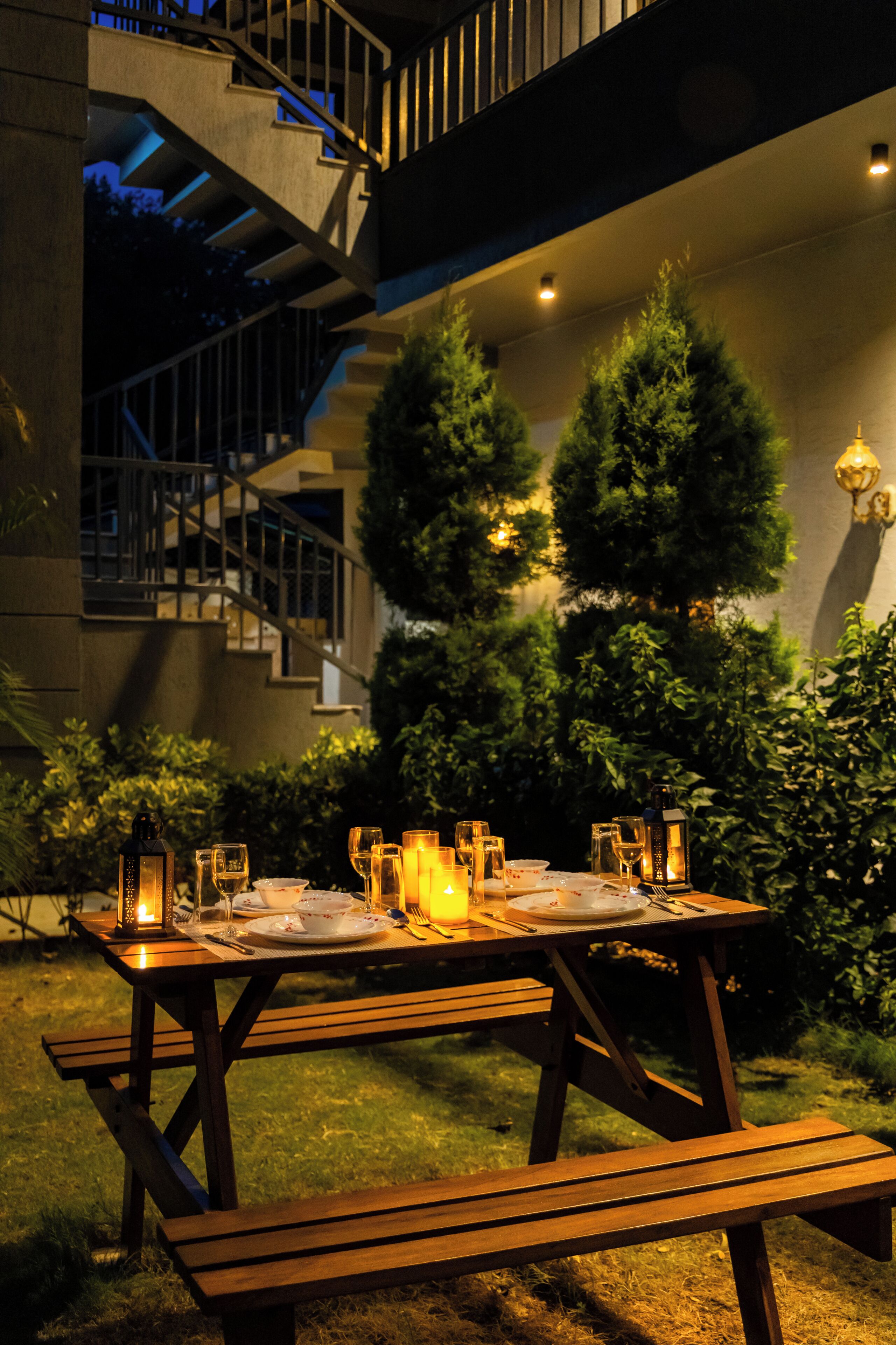 A wooden picnic table set up outdoors with candles, glasses, and cutlery.