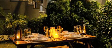A wooden picnic table set up outdoors with candles, glasses, and cutlery.