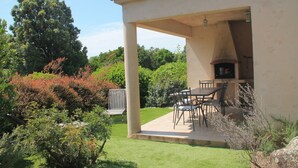 Eine überdachte Terrasse mit Tisch und Stühlen, umgeben von Pflanzen und Bäumen.