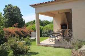 Eine überdachte Terrasse mit Tisch und Stühlen, umgeben von Pflanzen und Bäumen.