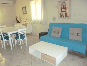 Ein Wohnzimmer mit einem blauen Sofa, zwei weißen Sesseln, einem weißen Tisch und einer kleinen weißen Kommode.